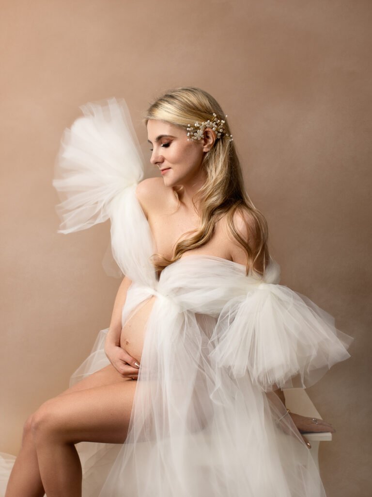 Kobieta w ciąży w białej sukni na beżowym tle - delikatna fotografia